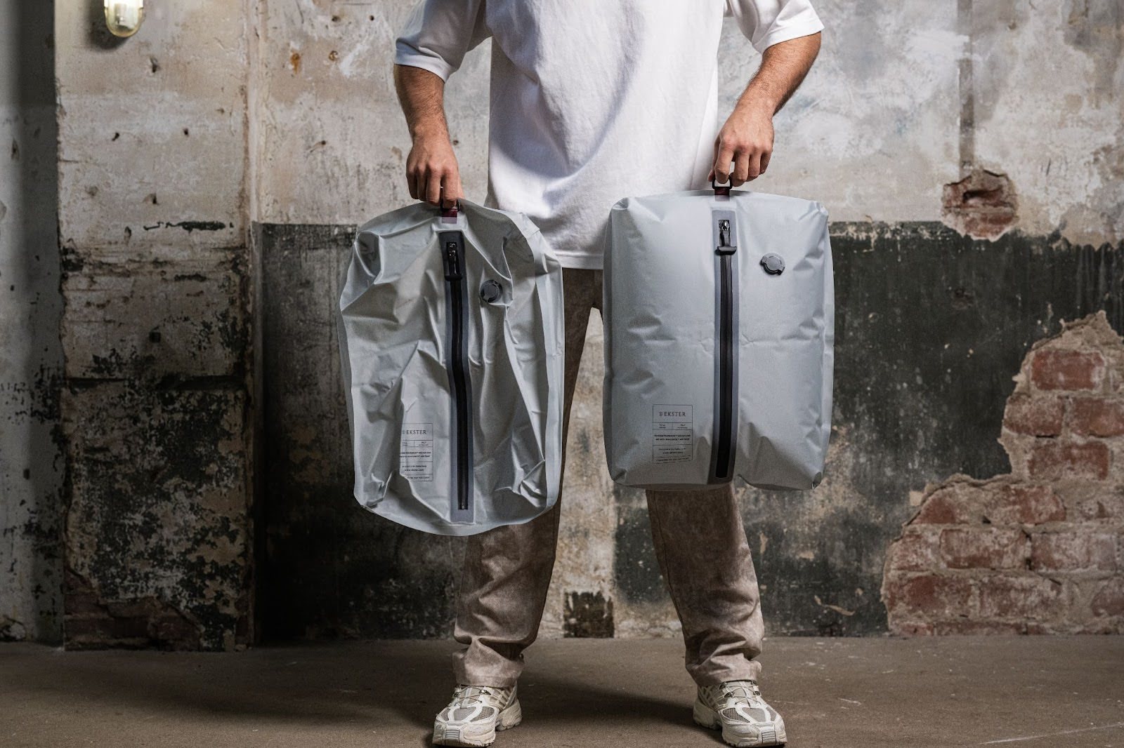 Person holding two grey waterproof bags with vertical black zippers, standing against a worn industrial-style wall.