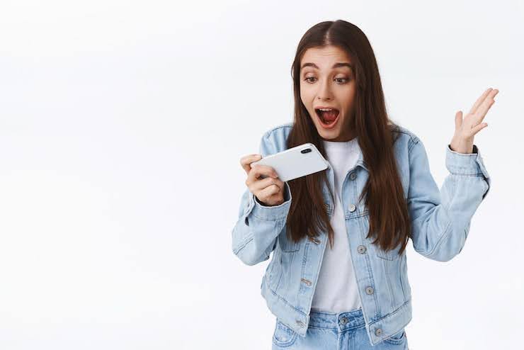 Impressed excited and thrilled young girl shouting at smartphone astonished raising hand up triumphing holding mobile phone horizontally stare screen amazed white background Technology concept