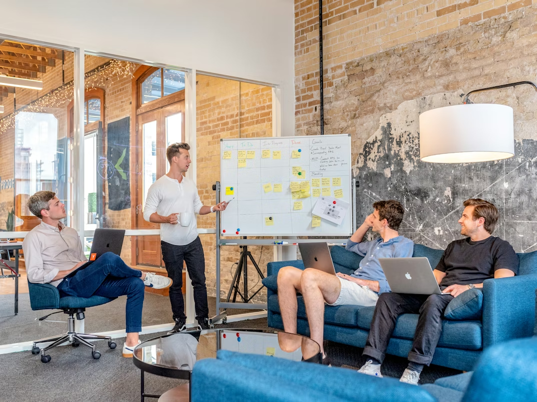 Four colleagues in a modern office with exposed brick, using laptops and discussing a whiteboard filled with sticky notes.