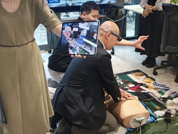 Person performs chest compressions on a CPR manikin while another holds a tablet showing the training, with an instructor nearby.