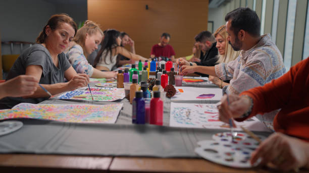 Group of adults sitting at a long table, painting with brushes and colorful paints during an art therapy session.
