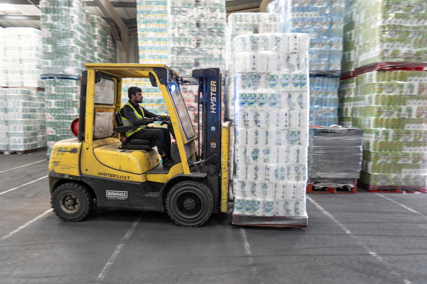Worker operating a yellow forklift carrying a tall load of packaged toilet paper.