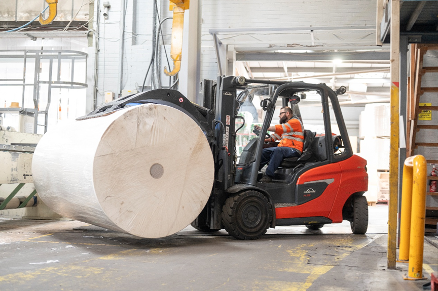 Worker in high-vis gear driving a red forklift moving a large industrial paper roll.