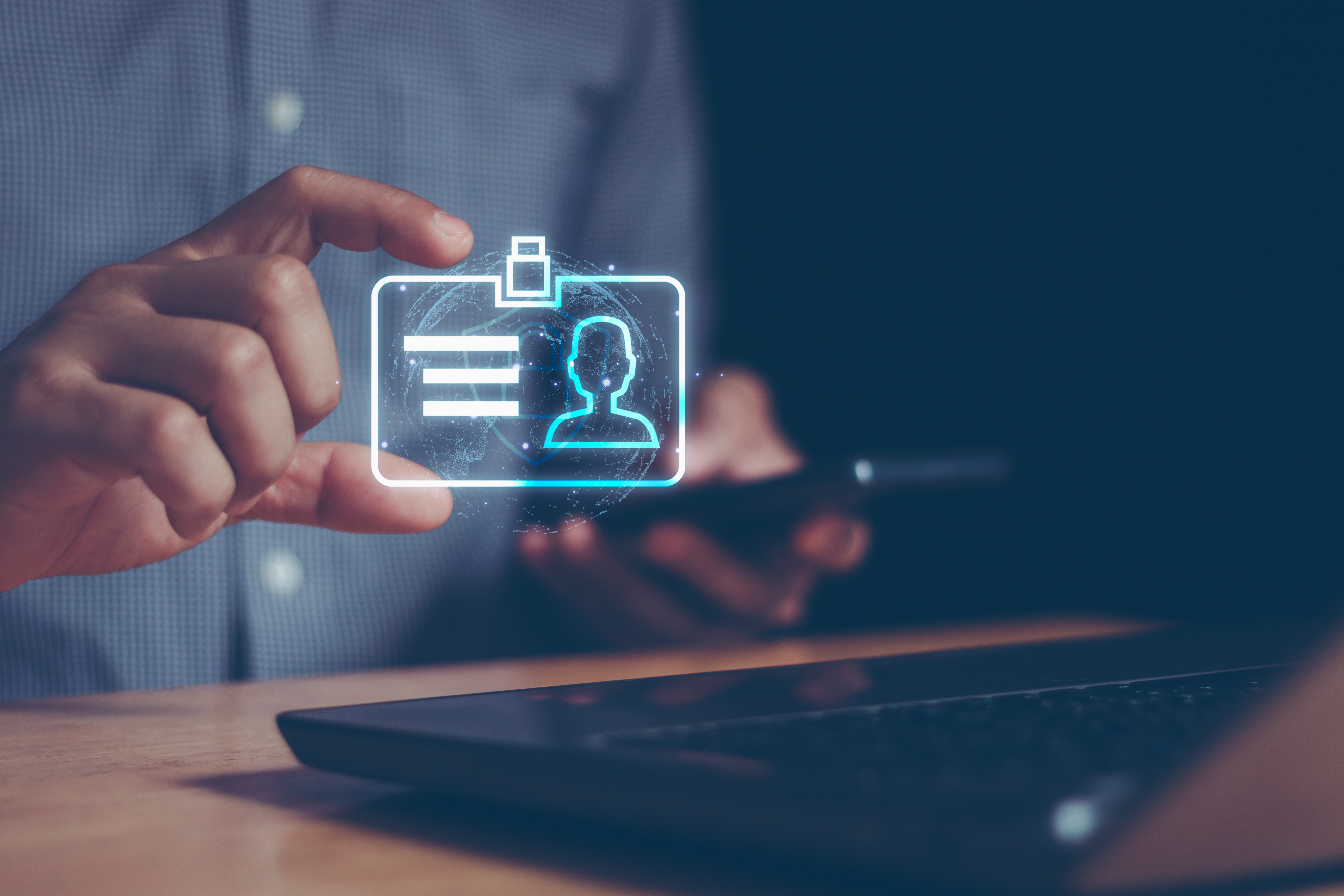 Person holding a smartphone while interacting with a glowing holographic ID card above a laptop, symbolizing digital identity.