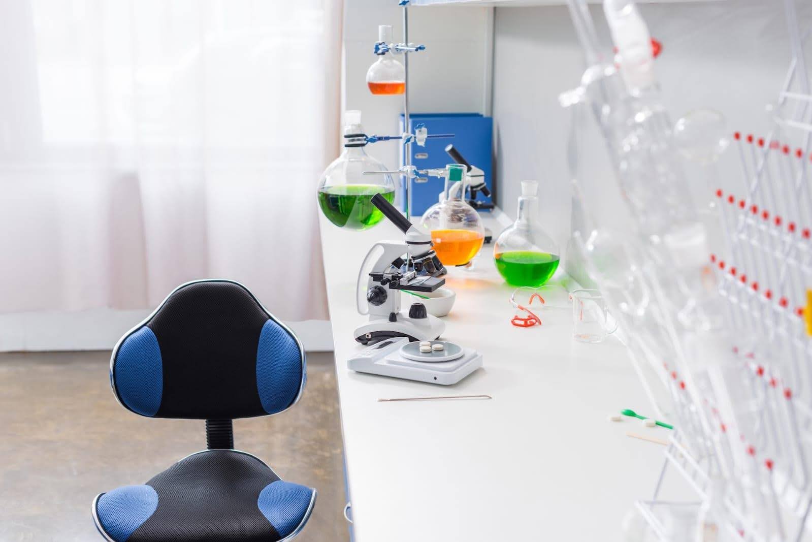 Modern lab setup with a microscope, colorful chemical flasks, and a black-blue chair beside a clean white workbench.