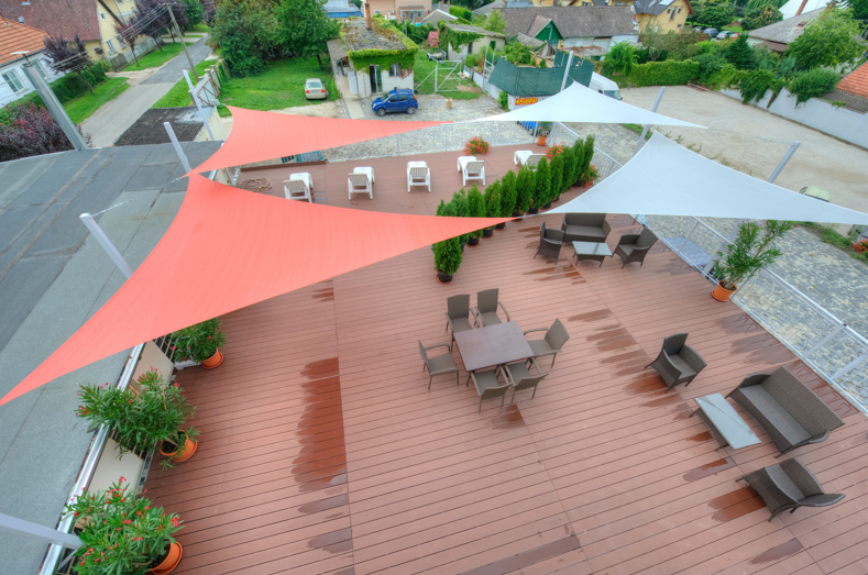 Deck area with seating, colorful shade sails, and greenery, set against a backdrop of a street and buildings.