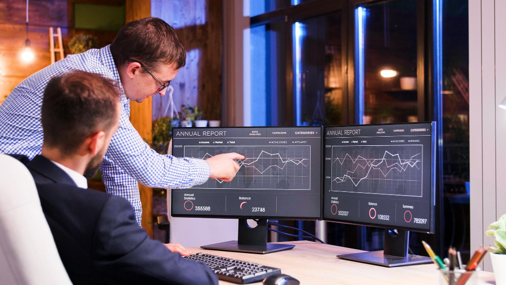 Two men are looking at and discussing data displayed on two computer monitors in an office.