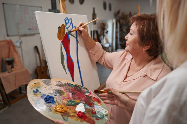 Older woman painting an abstract figure on a canvas while a person holds a palette of oil paints.