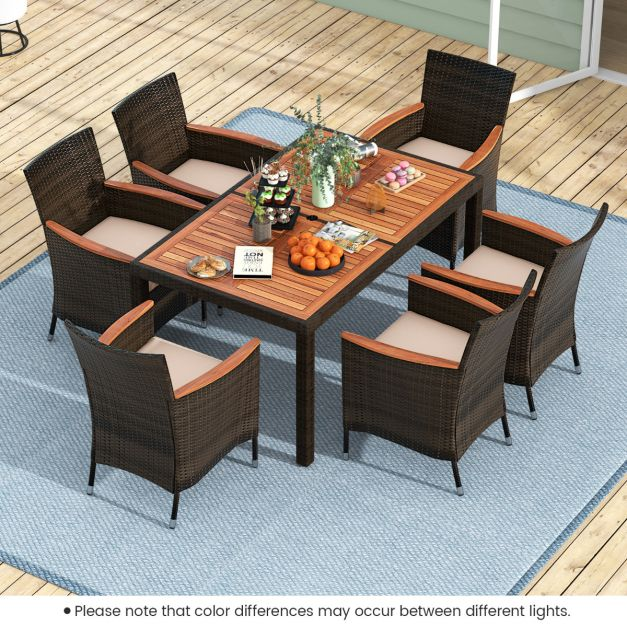 Outdoor dining set with a wooden table and six wicker chairs with beige cushions on a blue rug on a deck.