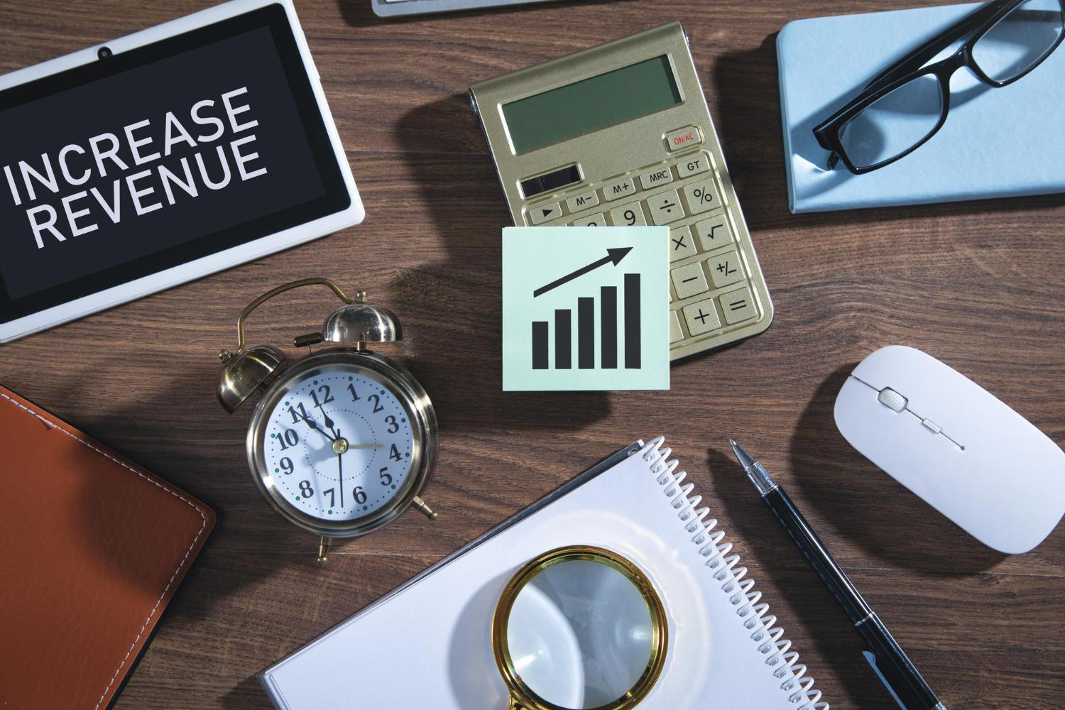 Overhead shot of a desk with an alarm clock, calculator, and sticky note showing a bar graph, and a tablet displaying "INCREASE REVENUE".