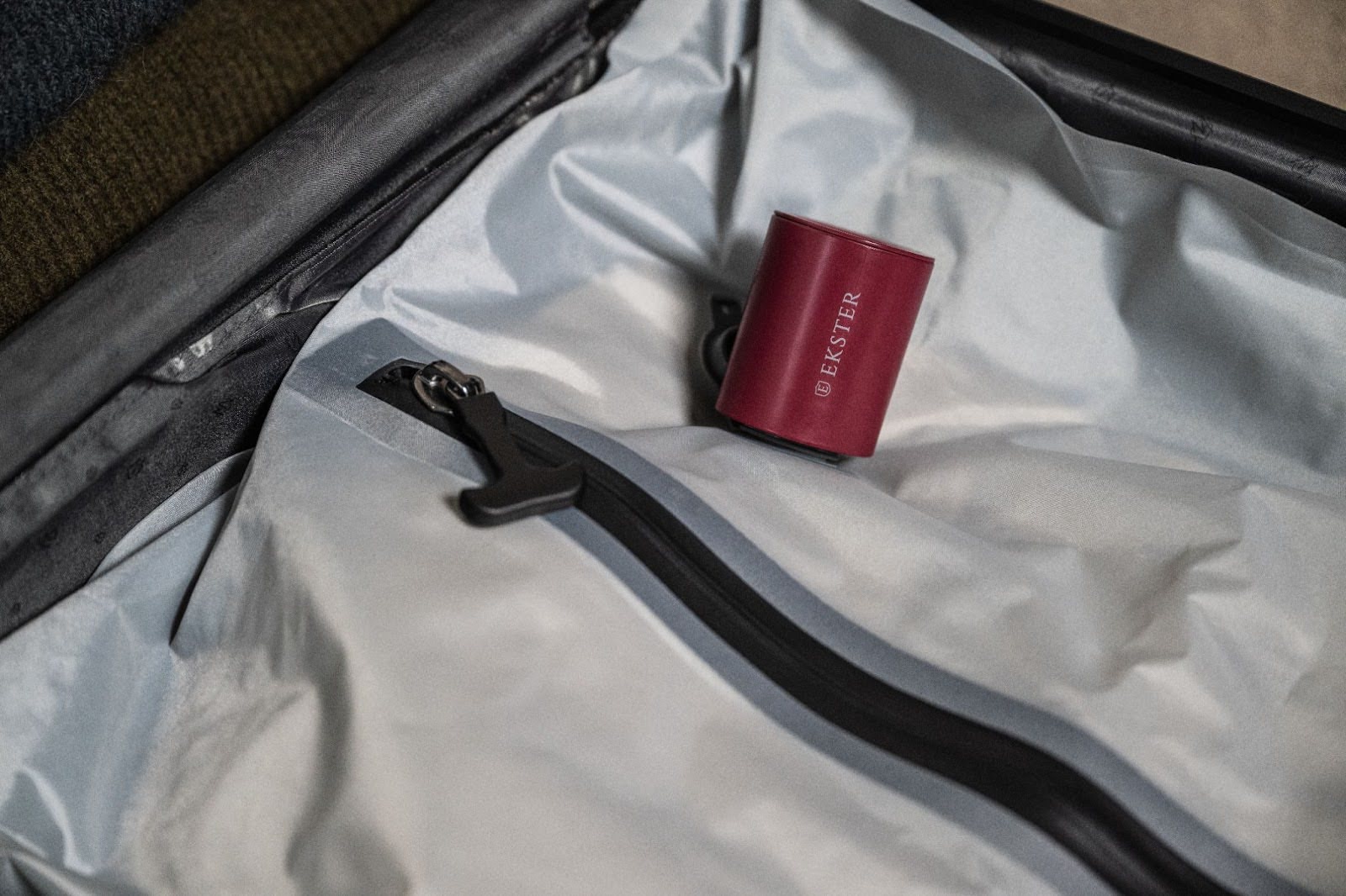 Interior of a suitcase with a zipper pocket, containing a small red cylindrical object labeled 'Ekster.'