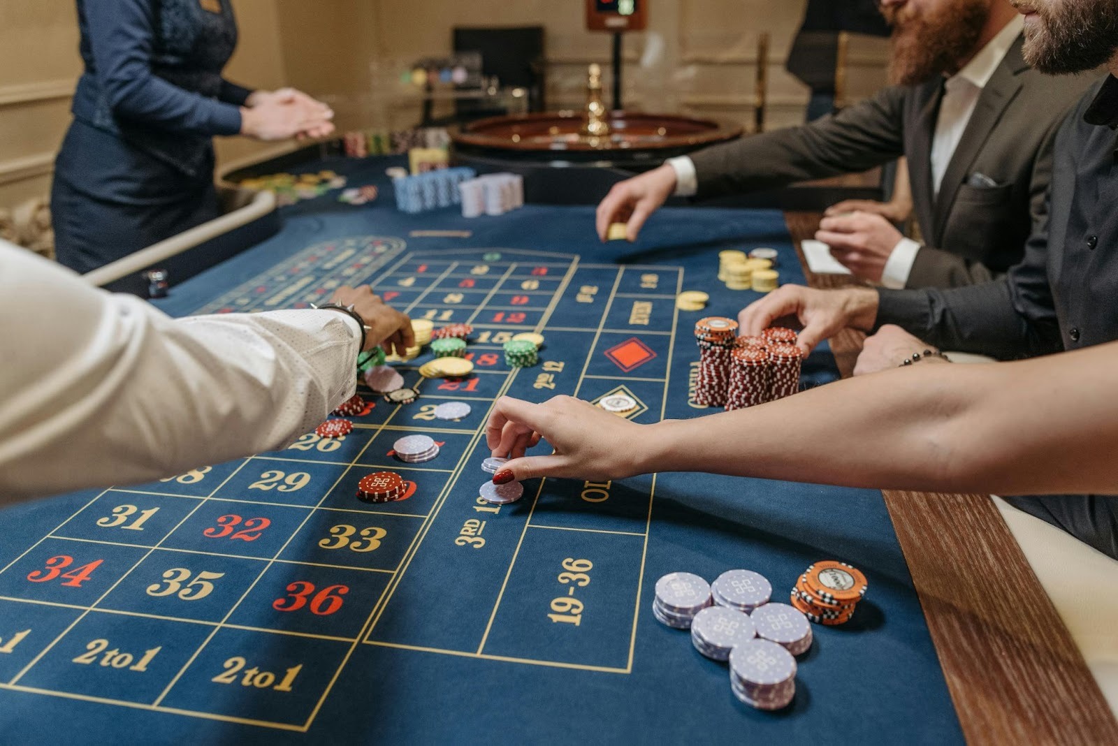 People placing bets with colorful chips on a blue roulette table in a casino.
