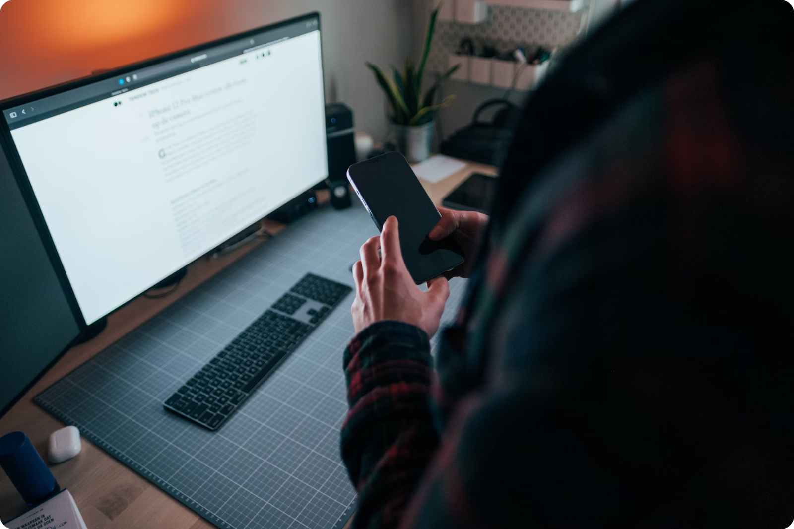 Person in a plaid shirt using a smartphone at a desk with a large computer monitor displaying an article.