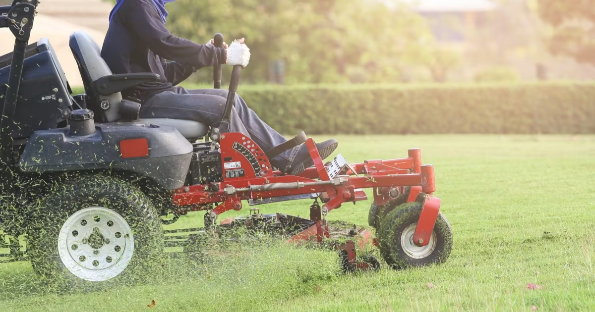 Person operating a ride-on lawnmower, cutting grass on a large sunny lawn.