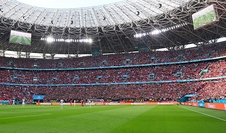 Crowded stadium with fans watching a live soccer match under a modern roof, with large screens showing the game.