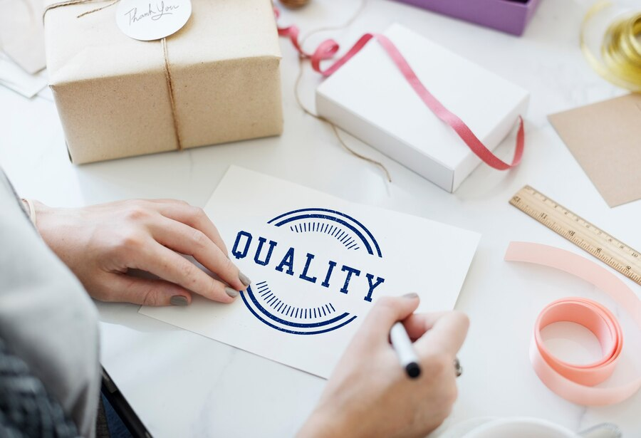 A person writing on a card labeled 'QUALITY' amidst gift-wrapping supplies, including ribbons, a gift box, and a ruler