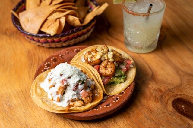A plate of two tacos is on a wooden table next to a basket of tortilla chips and a drink with a salted rim.