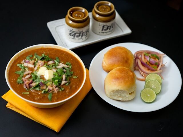 A bowl of pav bhaji with a pat of butter, alongside two buns, sliced onions, and lime wedges on a plate.