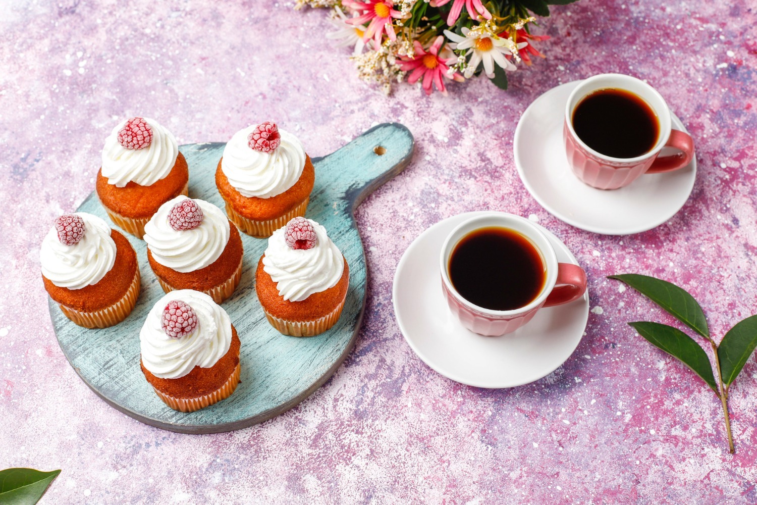Seven cupcakes with white frosting and raspberries on a cutting board, alongside two cups of black coffee.