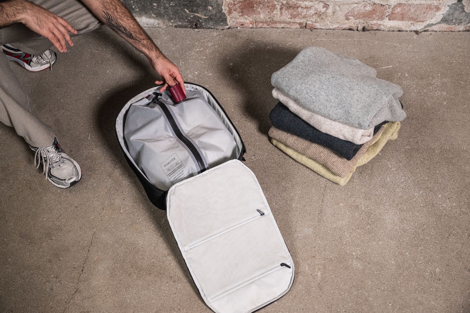 Person placing a small red cylindrical object into an open backpack with a white interior, labeled 'SETTER,' folded sweaters nearby.
