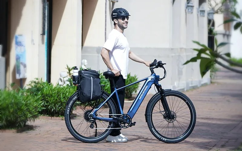 Smiling man wearing a helmet and sunglasses standing next to a blue electric bike with a rack bag on a sunny sidewalk.