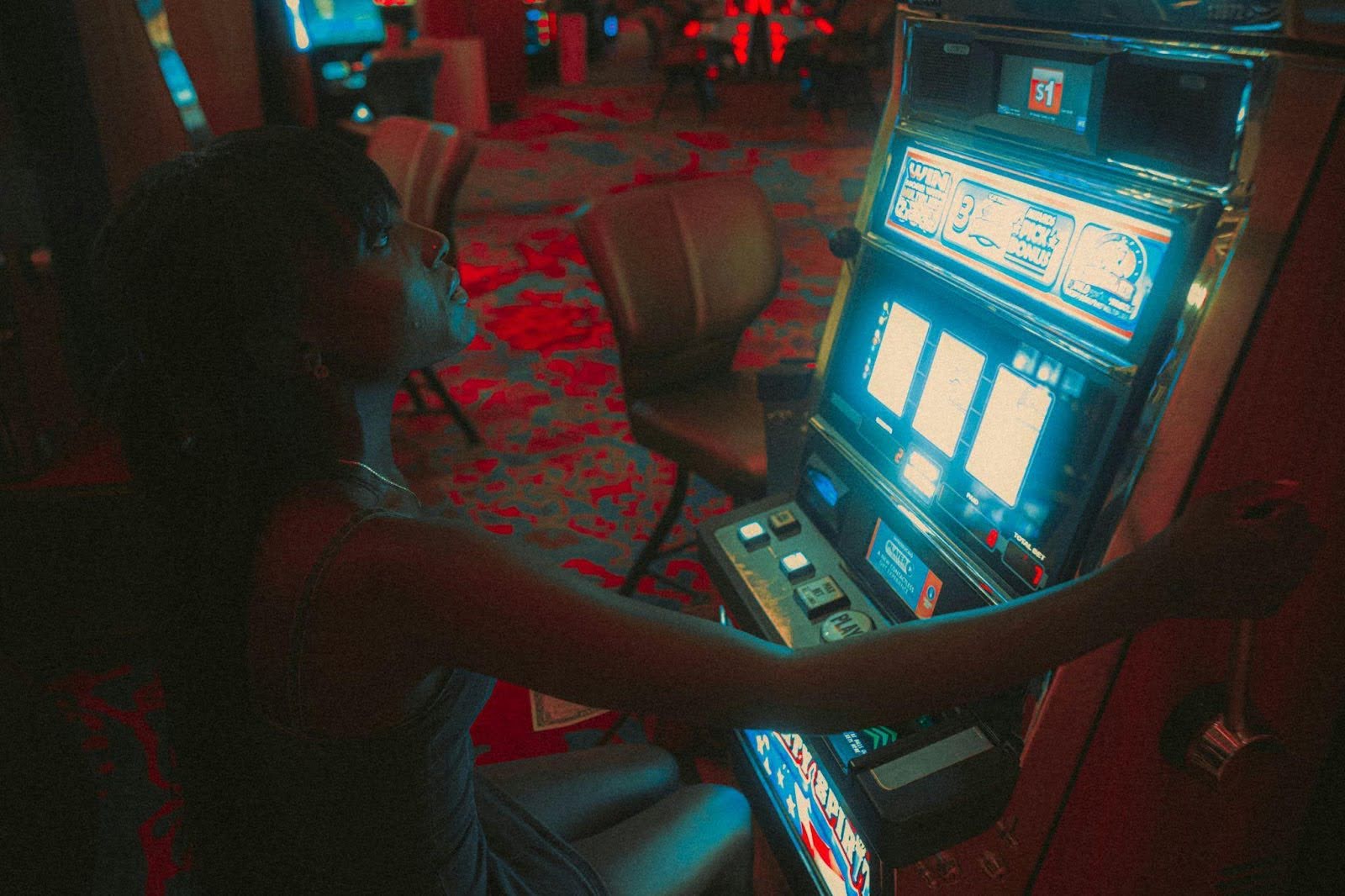 Person seated at a brightly lit slot machine in a casino, with patterned carpet and other machines in the background.