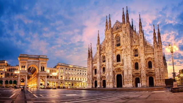 The Milan Cathedral (Duomo di Milano), a massive Gothic cathedral with intricate spires and statues, standing prominently in a large, open square at dusk.