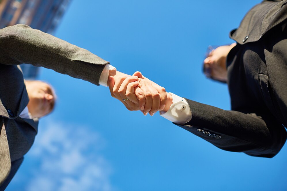 Two professionals in formal attire shaking hands, viewed from below against a bright, partly cloudy sky