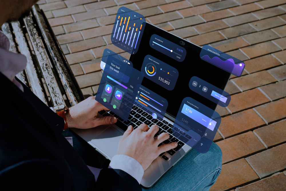  A person is sitting outside and using a laptop with transparent, holographic data and financial dashboards floating above the screen.