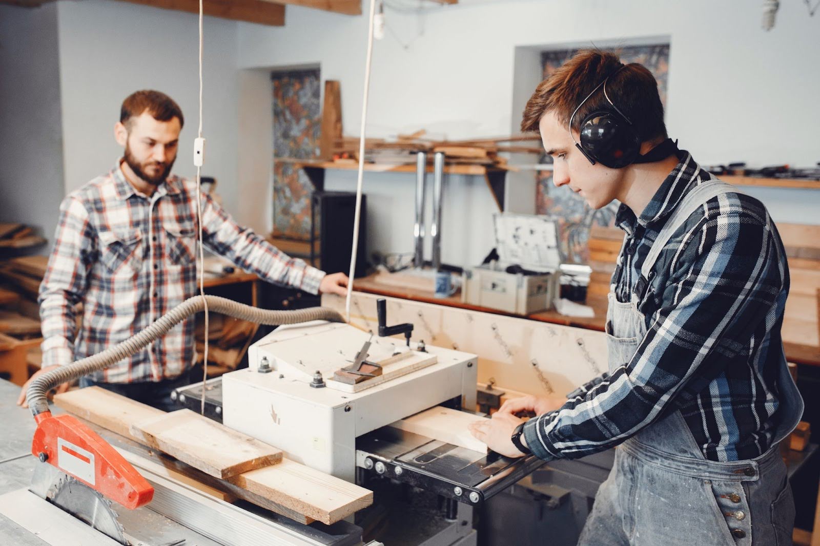 Two people in a woodworking shop, one using a table saw while the other observes, surrounded by tools and stacked wood.