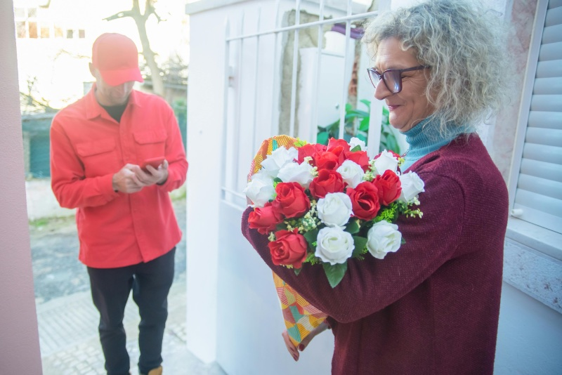 Woman accepting a bouquet of red and white roses from a delivery person in a red shirt who is checking his phone.