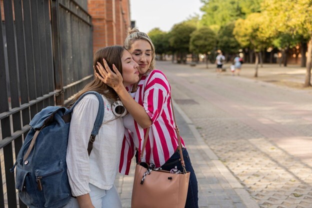 Woman embracing a child with a backpack on a sidewalk next to a fence.