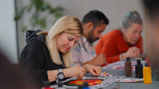Woman focused on painting a colorful design during an art class with other adults in the background.