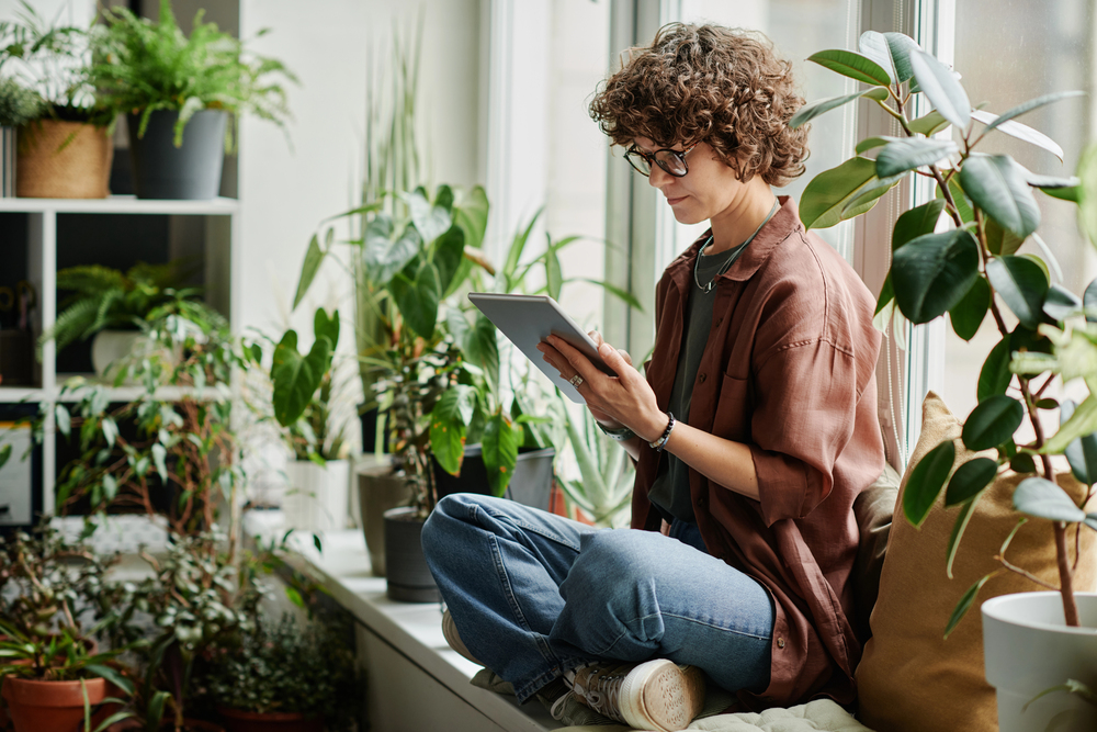 Young brunette woman with digital tablet sitting on windowsill