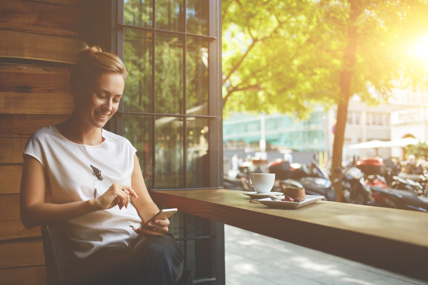 Smiling woman sitting in a cafe, looking at her phone with a cup of coffee and dessert on the table, sunlight streaming through the window.