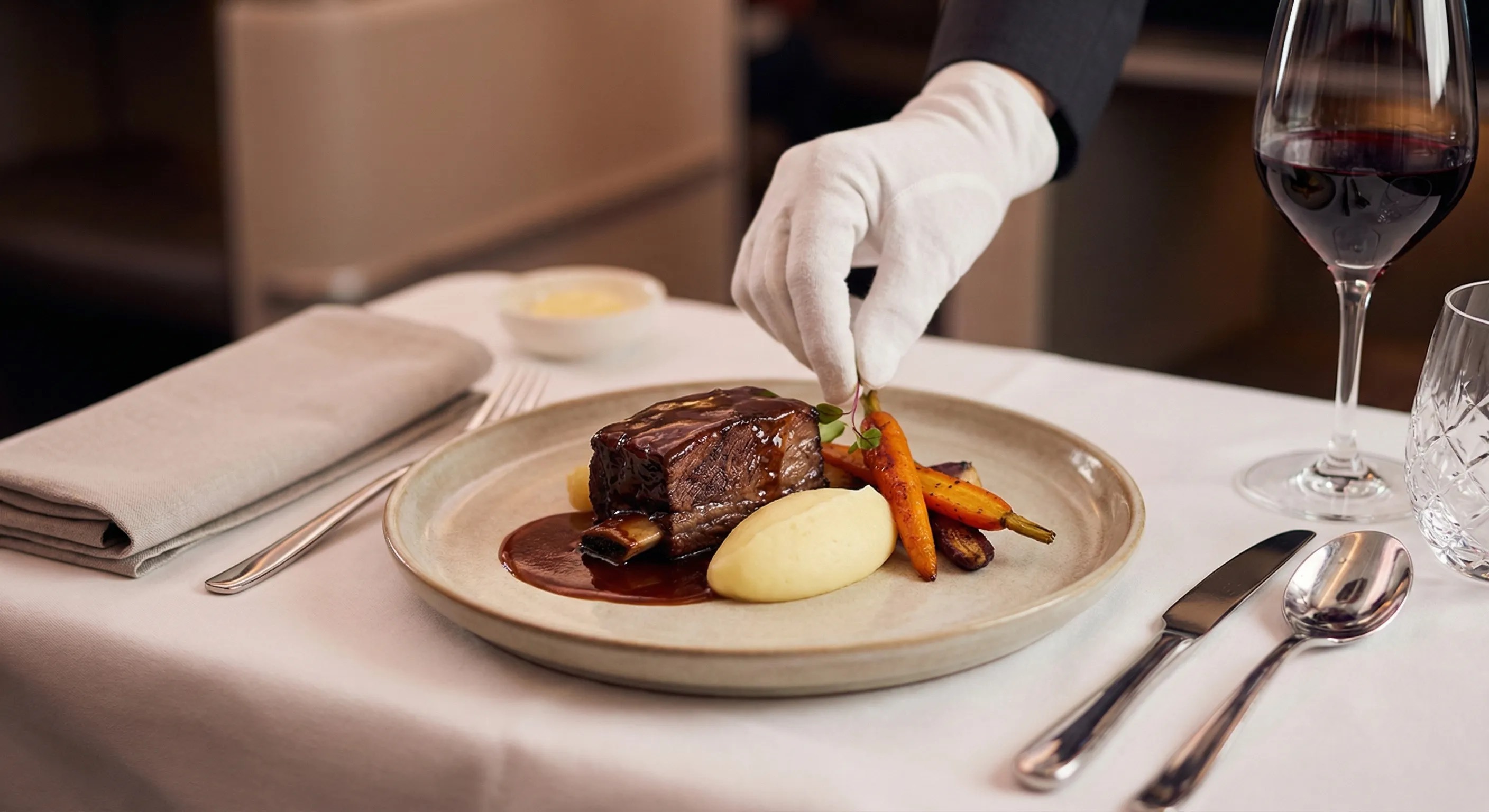 A close-up of a gourmet meal being served by a flight attendant with a white glove in a business class cabin, featuring braised beef and a glass of red wine.