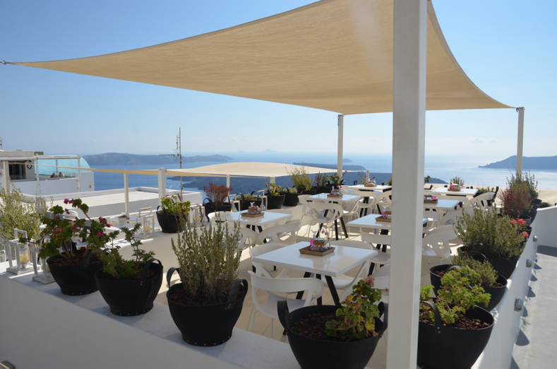"Outdoor seating area with white tables and chairs under a beige sunshade, surrounded by planters with greenery and flowers, overlooking a scenic coastal view.