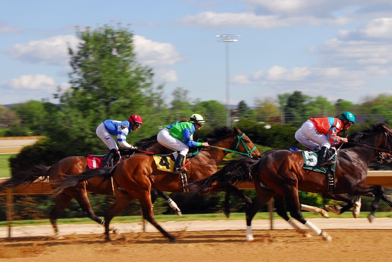 Three jockeys in colorful uniforms are riding horses at full speed on a dirt track during a competitive horse race, with a green landscape and trees in the background.