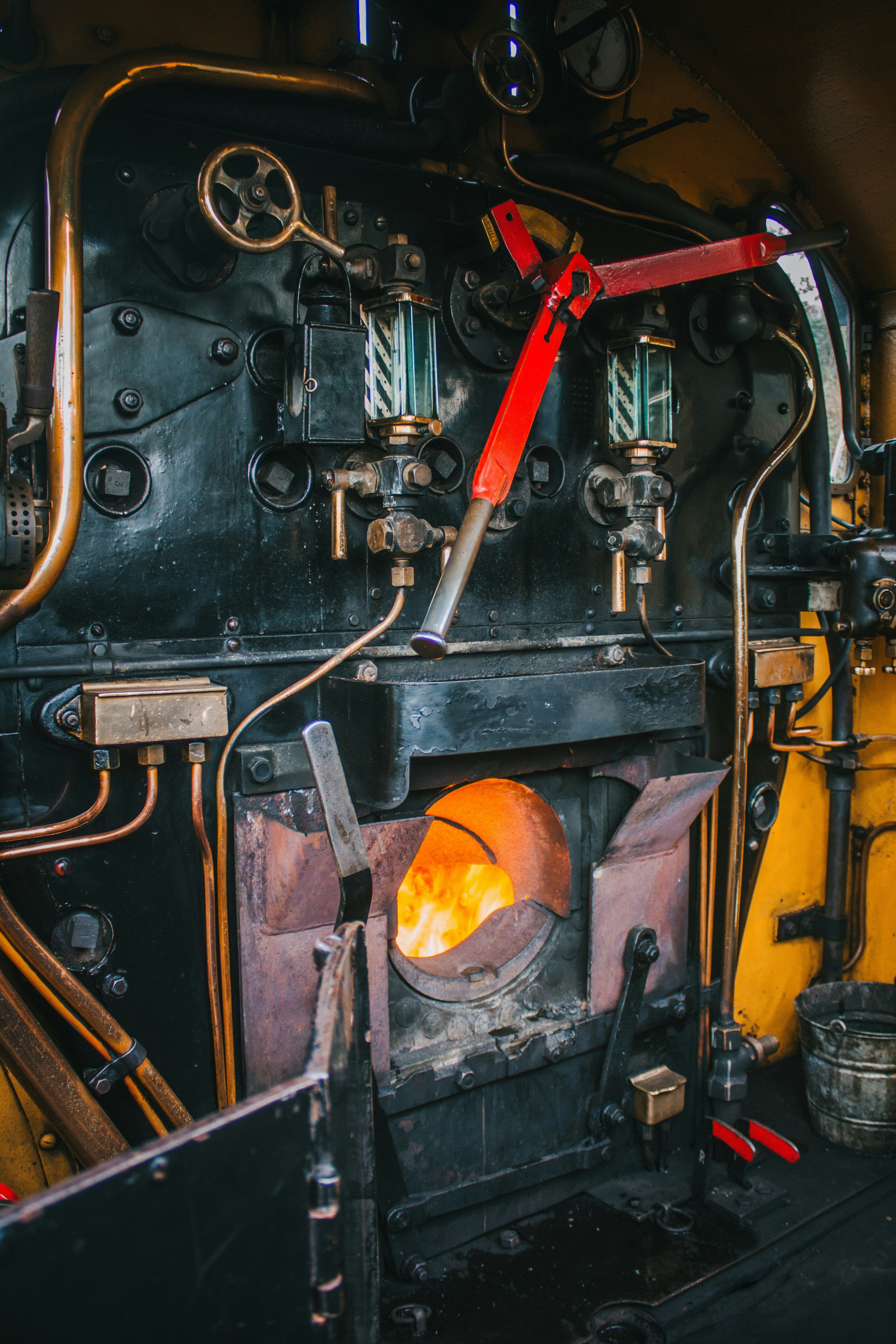 The interior of a vintage steam locomotive's boiler room, showing brass pipes, gauges, levers, and an open firebox with a glowing fire inside.