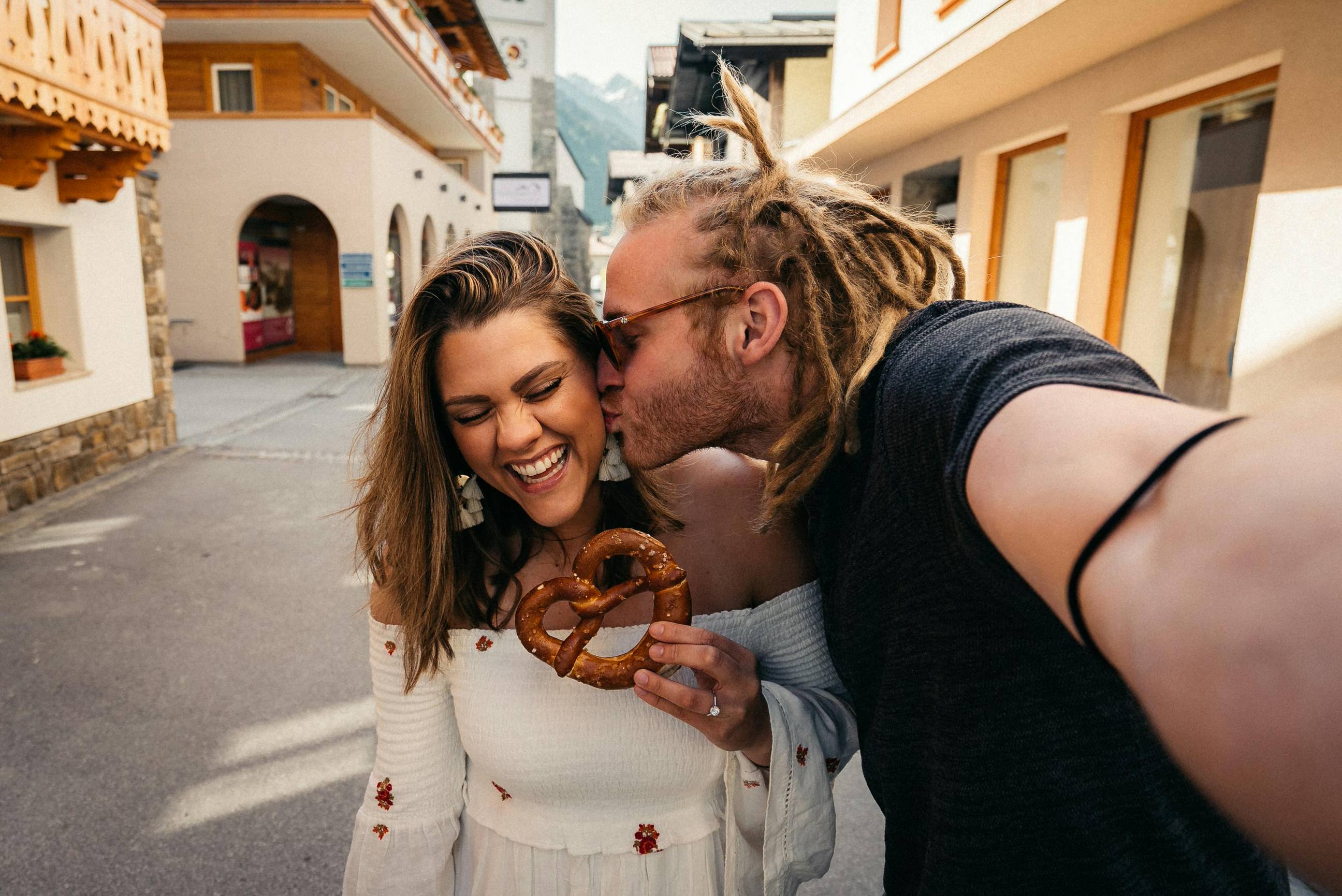 A couple taking a selfie, the man is kissing the woman's cheek while the woman is smiling and holding a pretzel