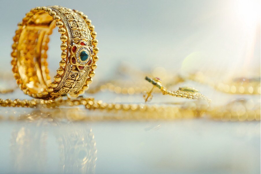 The Value of Artisanal Gold Jewellery - a photo close up of a decorative gold ring with green, clear and white stones in focus on a reflective white surface surrounded by other gold jewellery out of focus