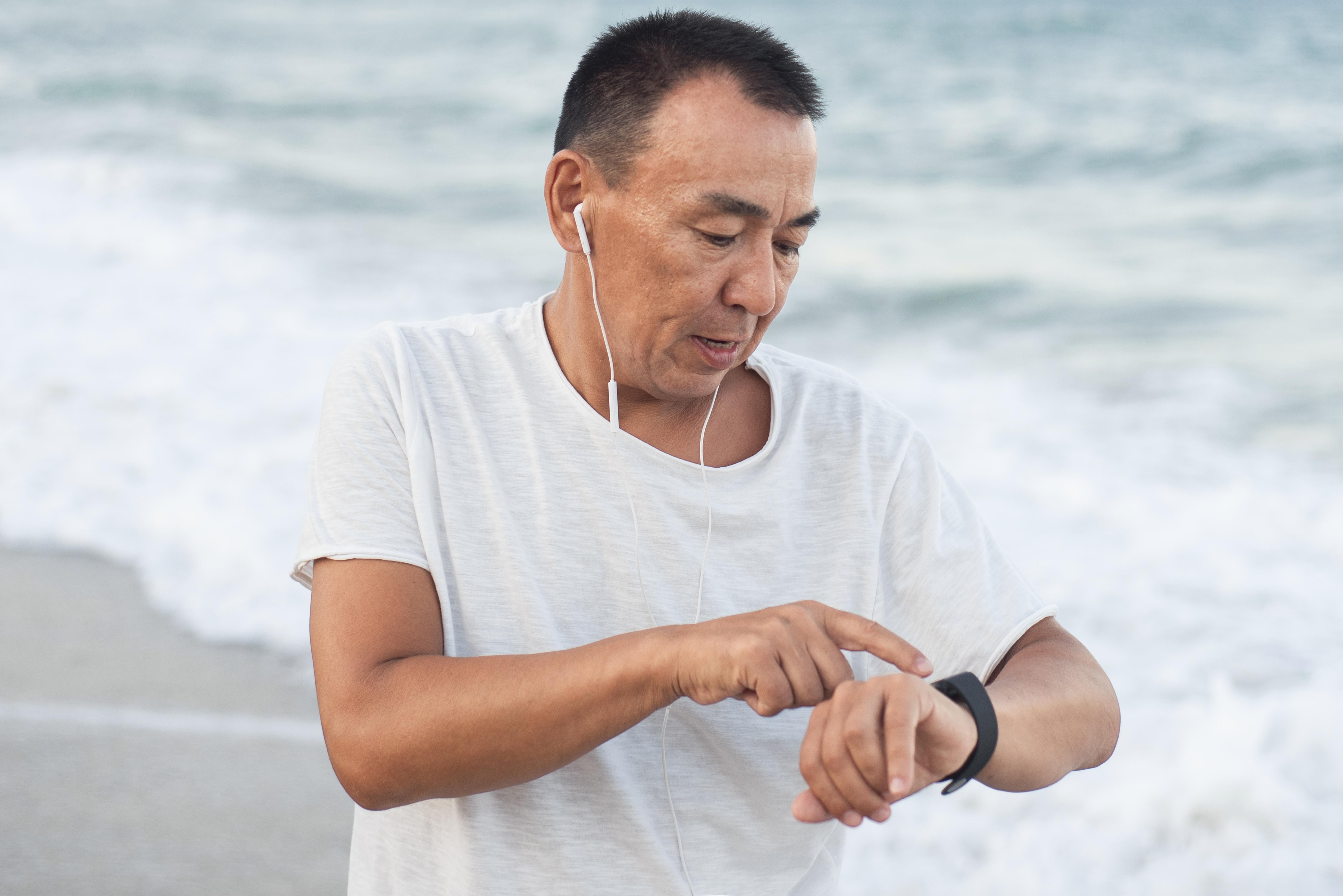 Person on a beach wearing earphones and pointing at a smartwatch, with ocean waves in the background.