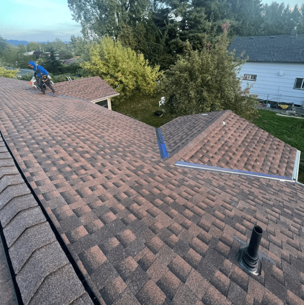 Person working on a brown shingled rooftop with tools nearby, surrounded by trees and neighboring houses
