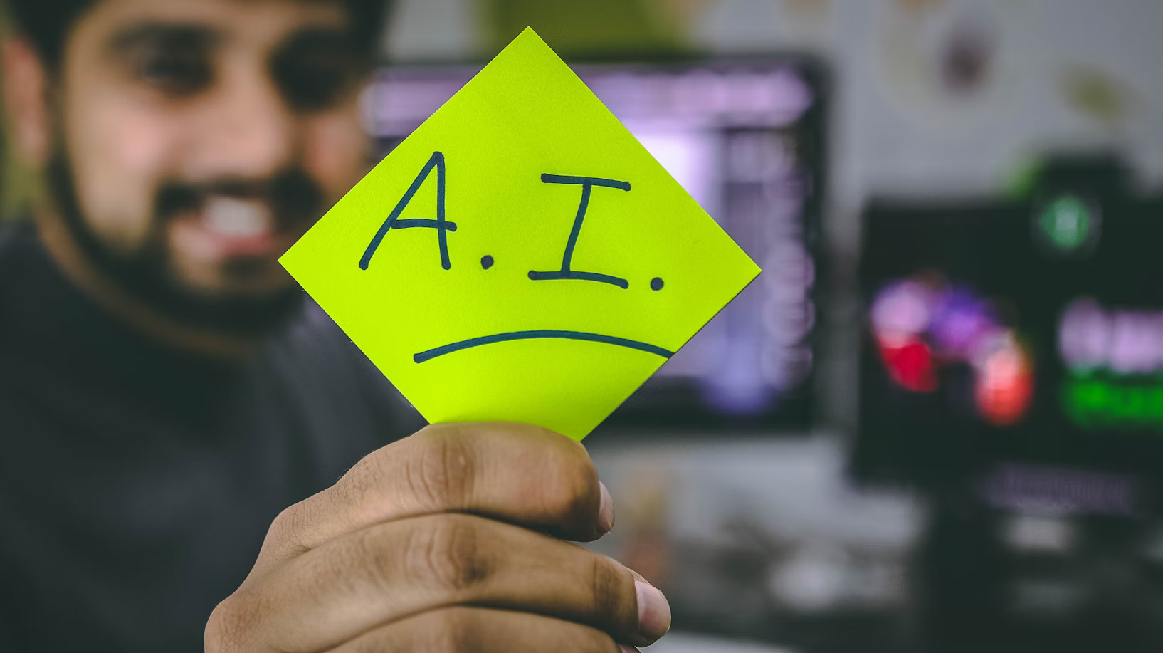 Smiling person holding a neon green note with “A.I.” and a frown drawn on it, in a tech workspace.