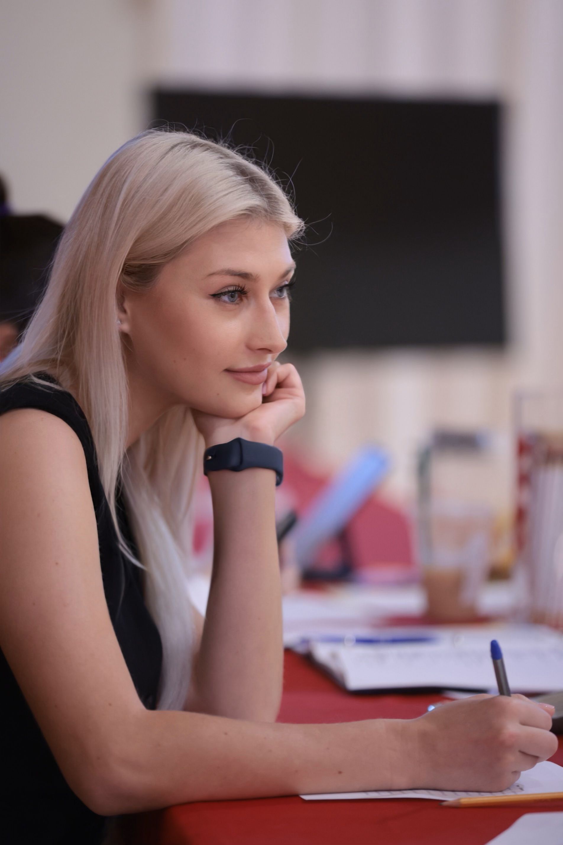 Person with long blonde hair writing in a notebook at a table, surrounded by papers and a drink.
