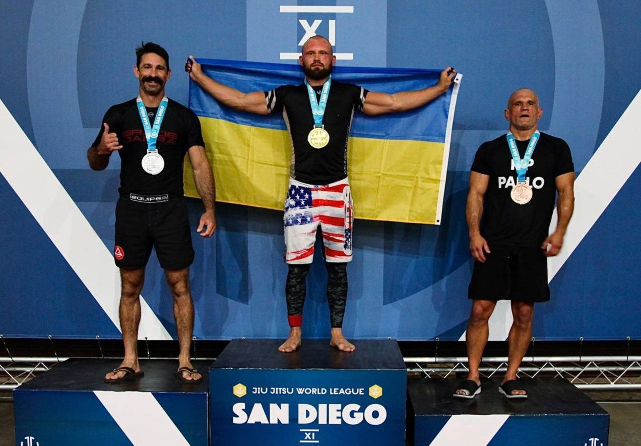 Three male athletes on a Jiu Jitsu World League podium in San Diego, with the gold medalist holding a Ukrainian flag.
