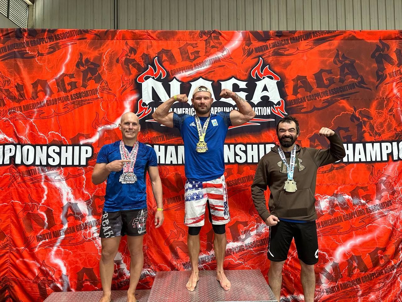Three male competitors posing with medals on the NAGA Championship podium, in front of a red event banner.