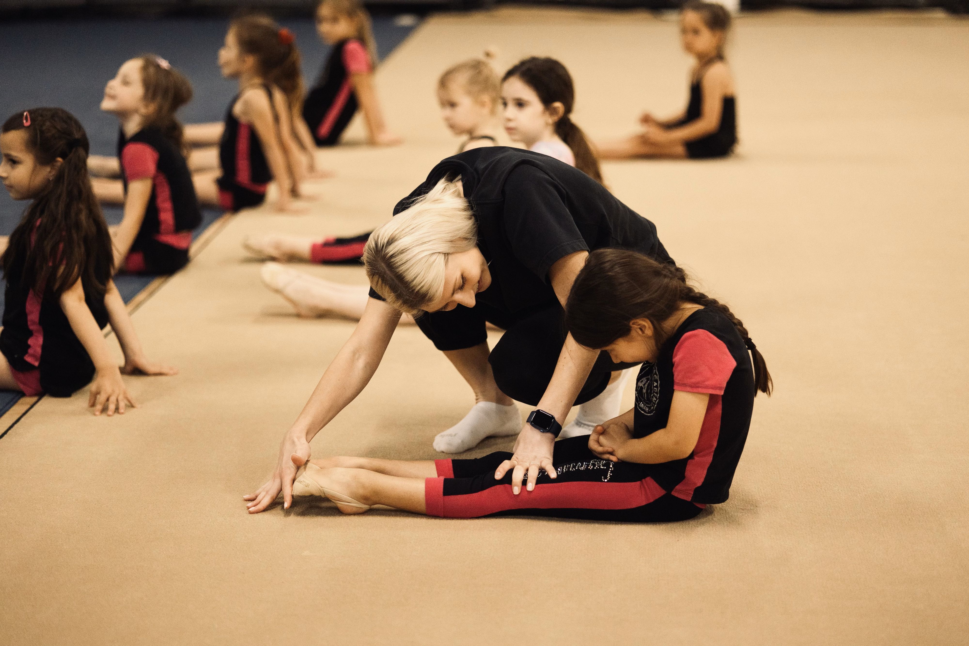 Gymnastics coach assisting a young girl with leg stretches as other children join the session in the background.
