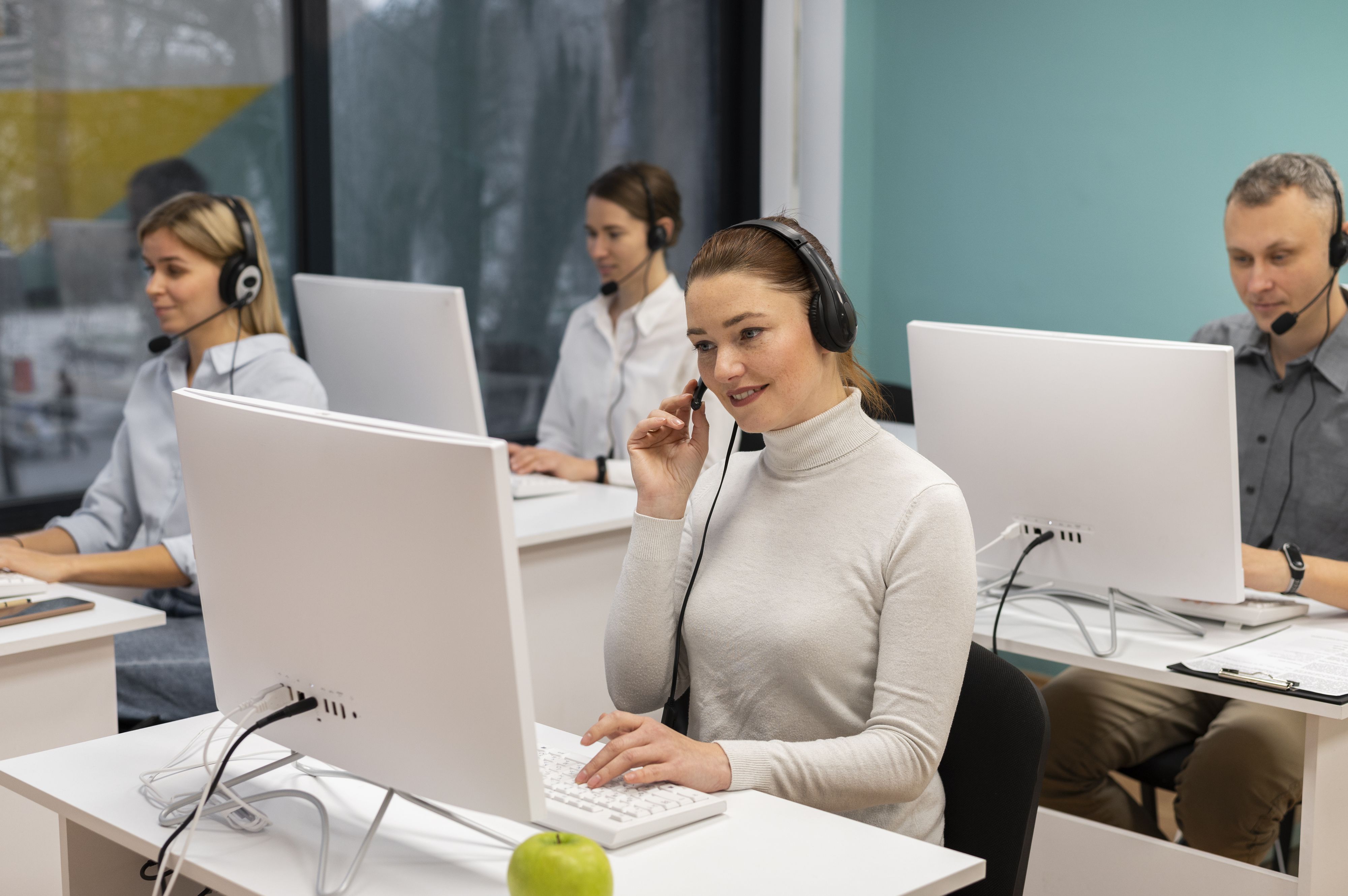 People wearing headsets working at computers in a modern office, with an apple on the desk in the foreground.