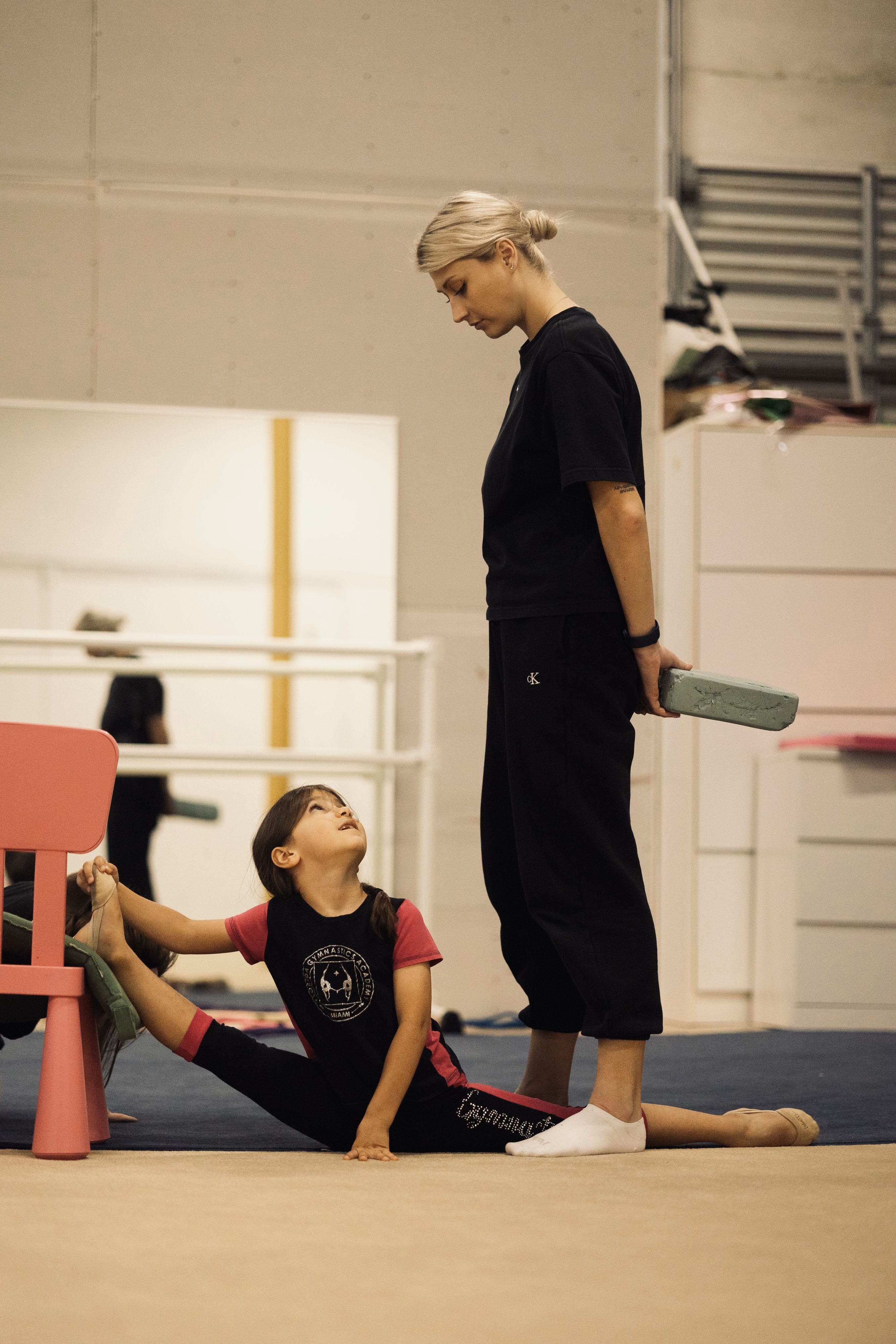 Young gymnast using a pink chair for support while stretching, guided by a coach holding a foam block in a gym setting.
